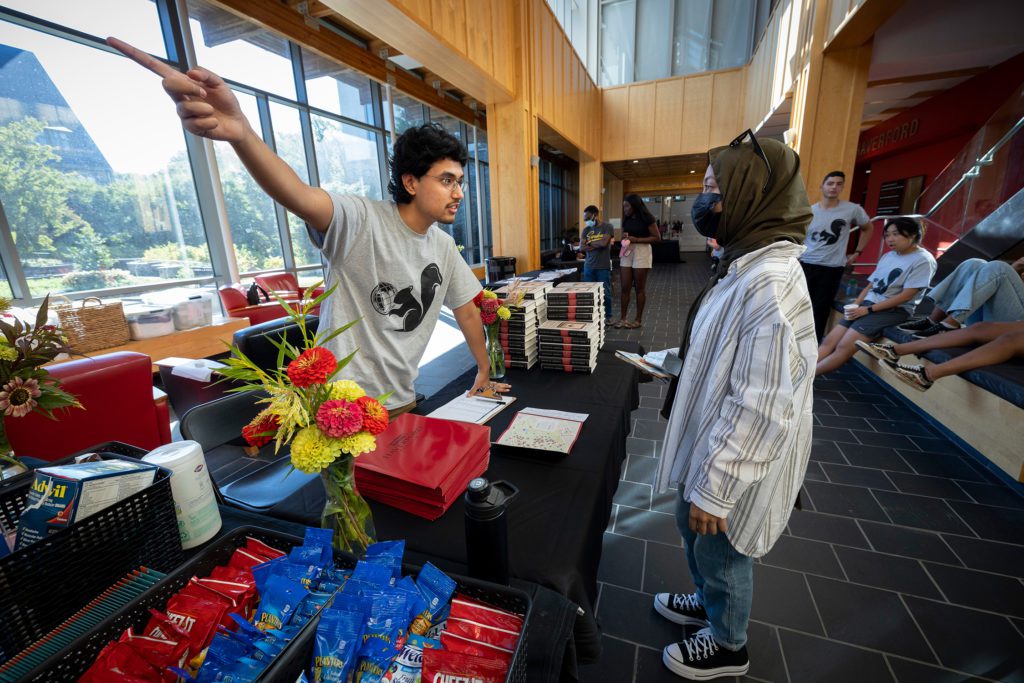 A student in a black squirrel t-shirt points and gives directions to a new student in a mask and a headscarf.