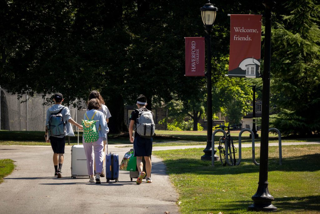 Four people photographed from behind wheel suitcases past a lamppost banner reading "Welcome, friends"
