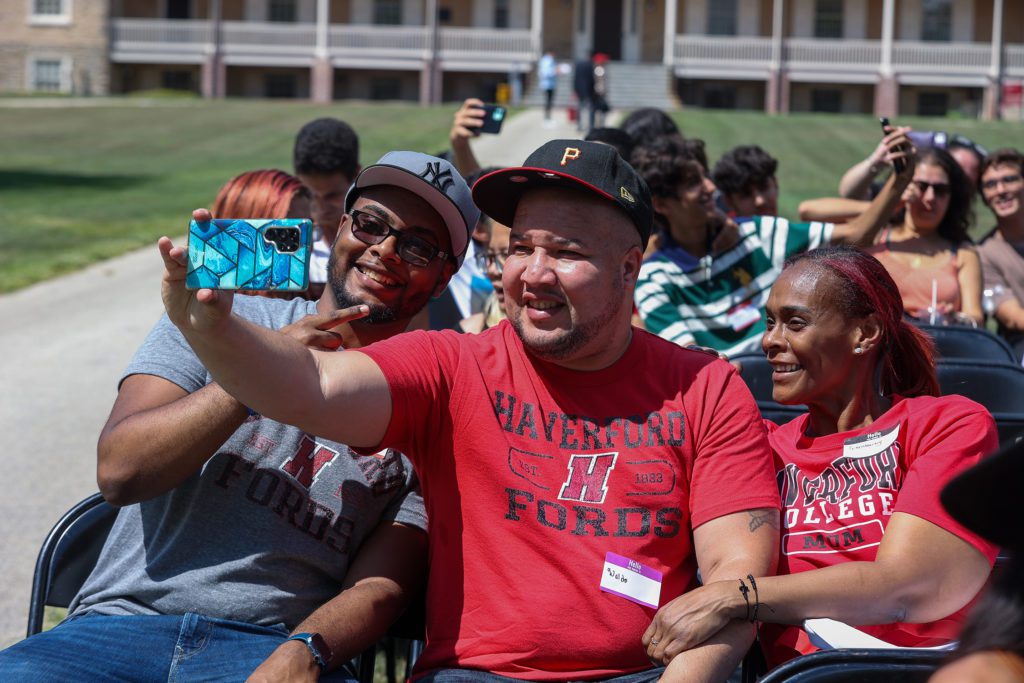 A student and his parents, all in different Haverford College t-shirts, take a selfie on Founders Green.