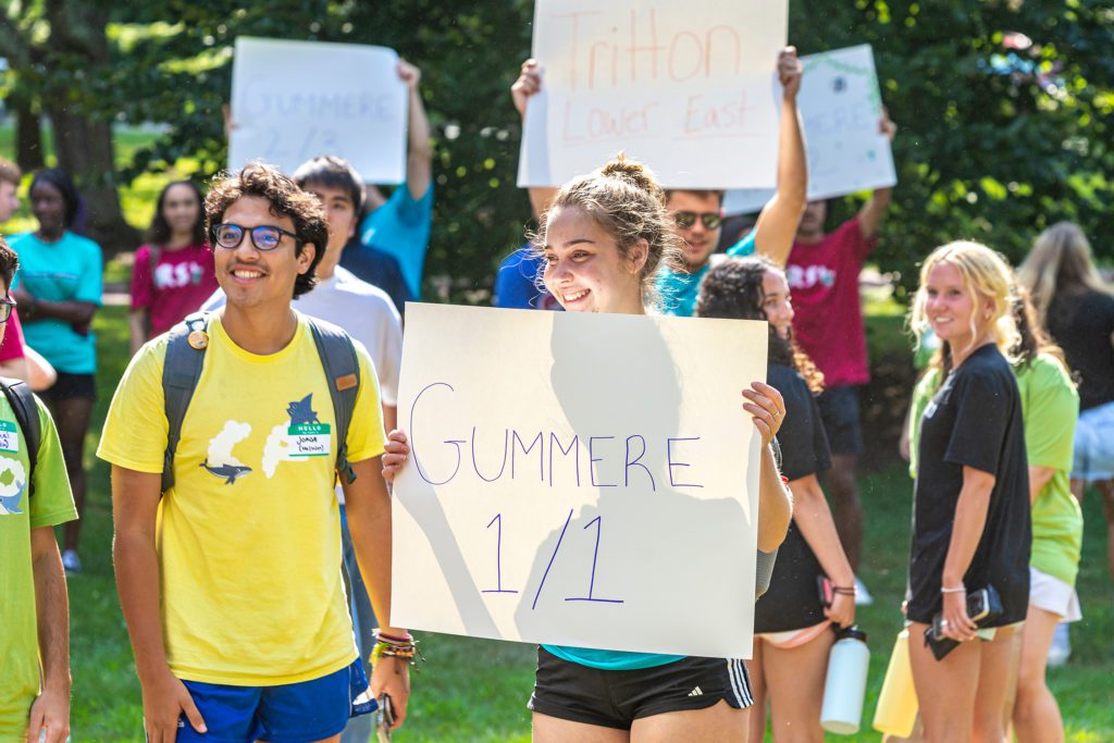 Customs teams hold up signs with different dorm names on it. In front is a girl enthusiastically holding a "Gummere 1/1" sign.
