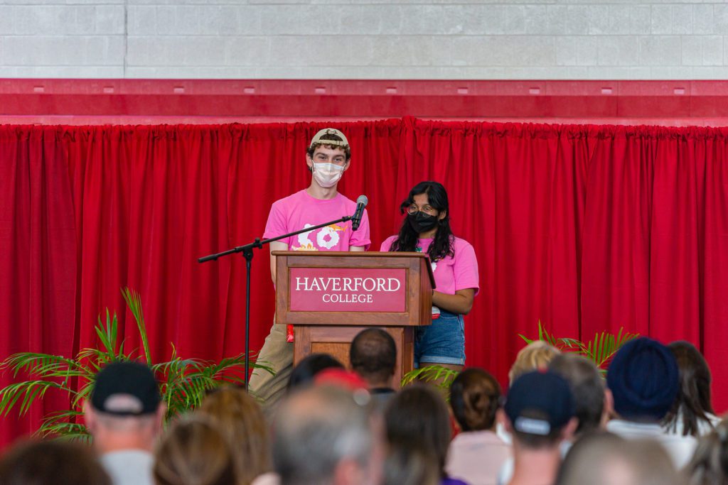 The two student Customs co-heads in pink t-shirts wear masks and speak at the podium.