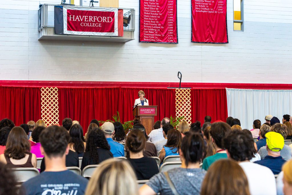 The president speaks at a podium under a Haverford College banner in the basketball gym in front of a crowd.