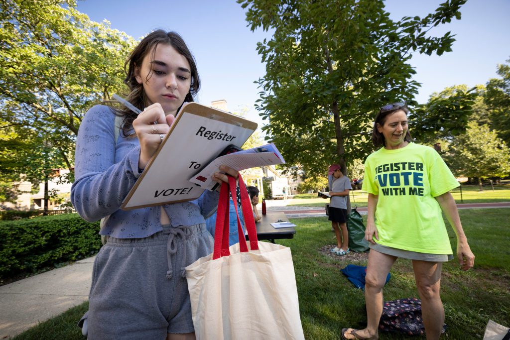 A new student registers to vote at orientation, standing next to a woman in a fluorescent yellow shirt that reads "register to vote with me"