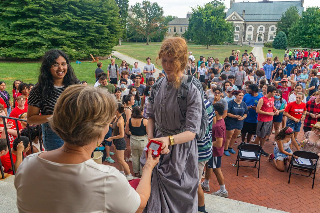 A student dressed in gray holds a tiny etch-a-sketch up to President Wendy Raymond to judge the drawing.