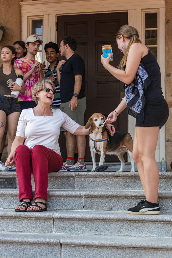 President Raymond sits on the steps of Founders with her beagle, who enjoys peanut butter from a spoon offered by a student.
