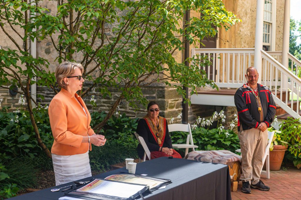 Wendy stands at a table, wearing sunglasses, speaking while members of the LNPA look on next to her.