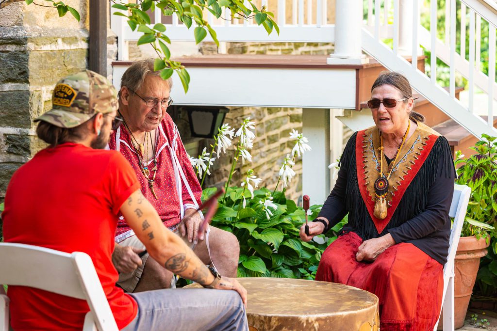 Three people sit around a large drum and bang it with mallets.