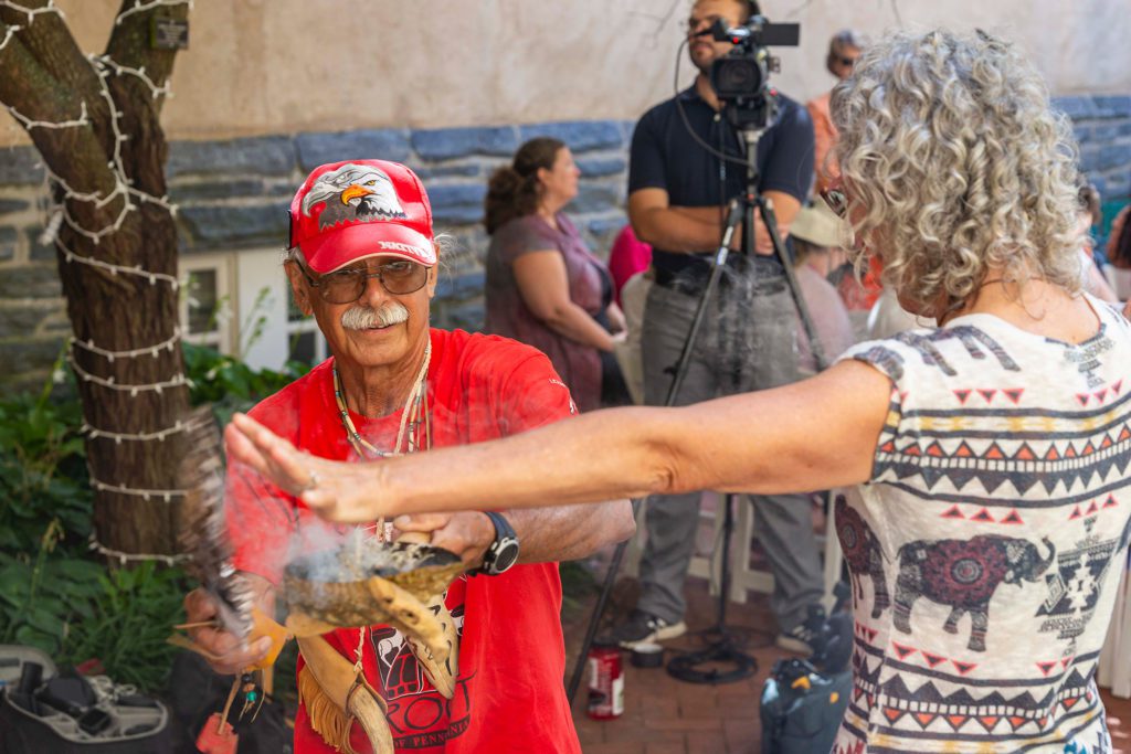 A member of the Nation encourages smoke from a clamshell bowl to cover a woman standing with her arms outstretched.