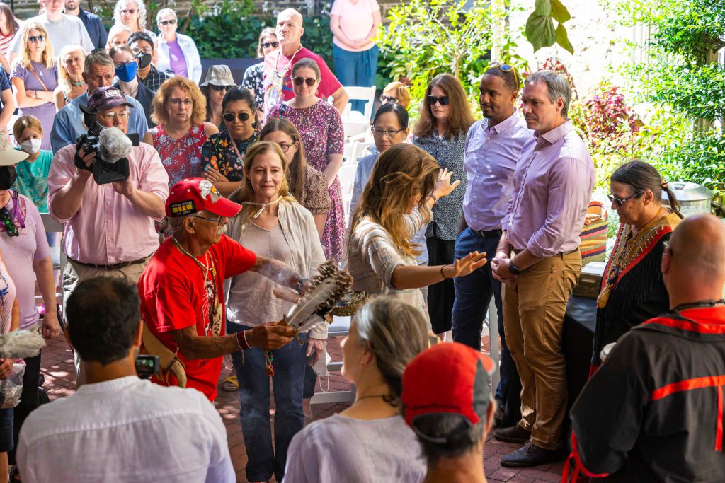 A crowd gathers around a person who has their arms out as a member of the LNPA waves smoke from a clamshell bowl over them with some feathers