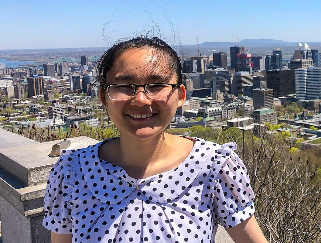 Lucy smiles wearing a polka dot blouse and glasses with a city skyline behind her