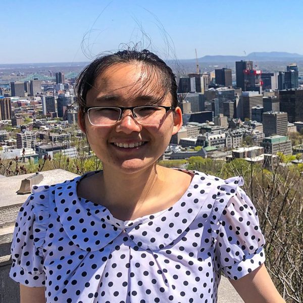 Lucy smiles wearing a polka dot blouse and glasses with a city skyline behind her