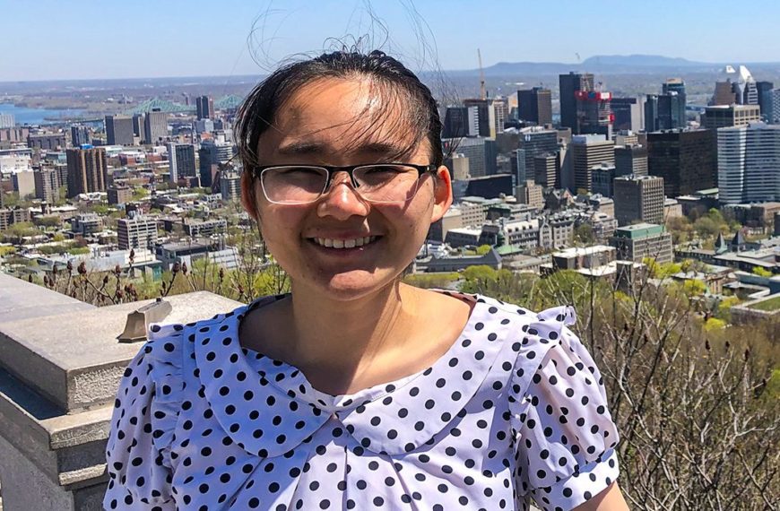 Lucy smiles wearing a polka dot blouse and glasses with a city skyline behind her