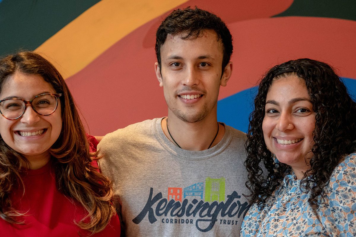 Ben stands between his two supervisors and in front of a rainbow mural
