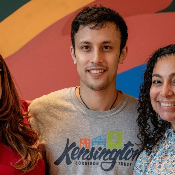 Ben stands between his two supervisors and in front of a rainbow mural