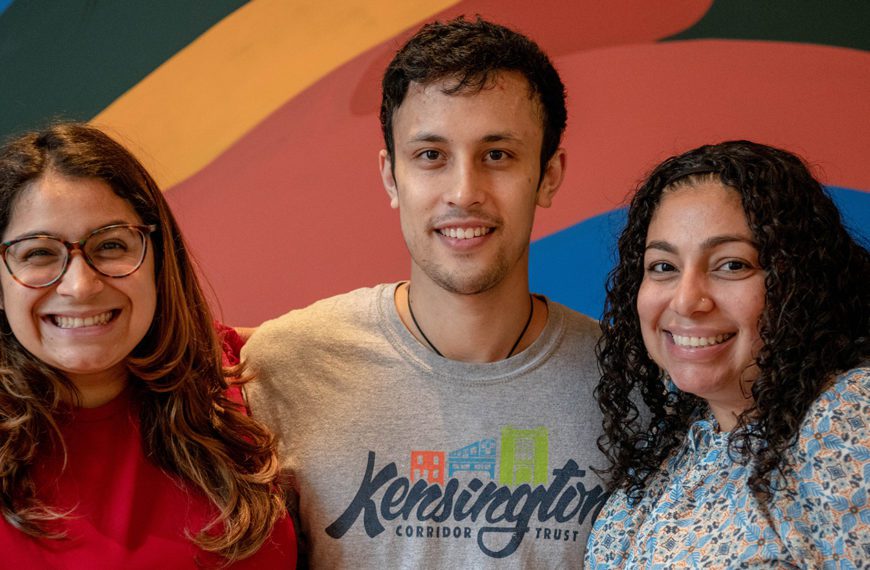 Ben stands between his two supervisors and in front of a rainbow mural