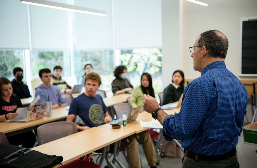 Richard Freedman stands at the front of class in. front of two rows of students seated at laptops.