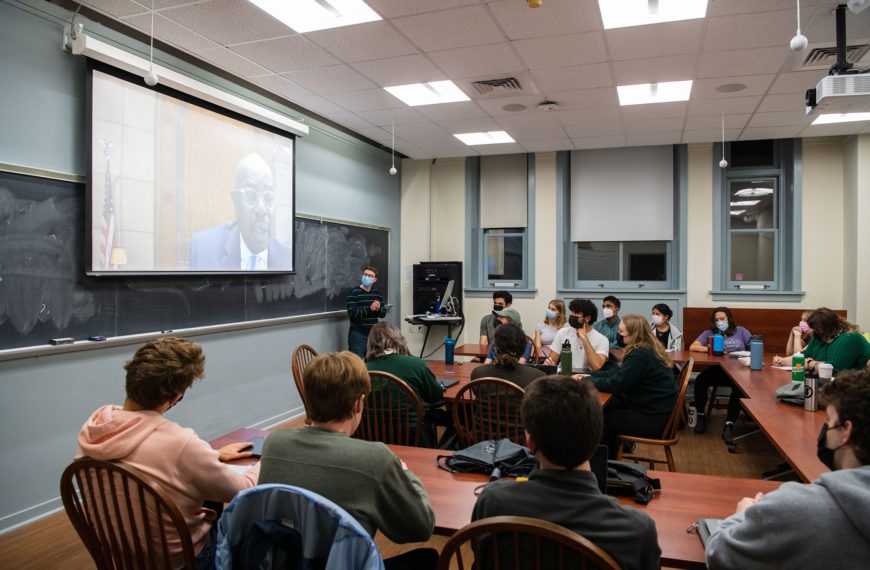 Students sit around tables in a seminar room and the judge's large face appears onscreen in front of them.