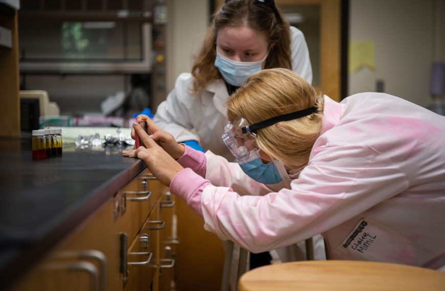 A student in a pink lab coat, goggles, and a mask leans over a counter and looks in a vial while another masked student in a lab coat looks on.