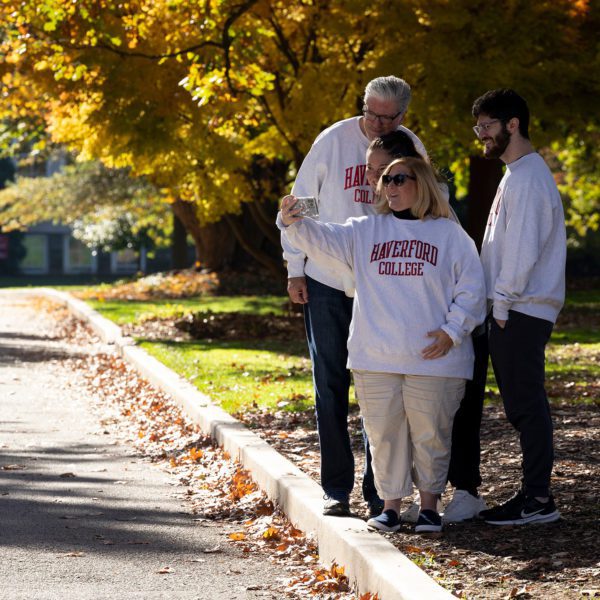 A family in matching Haverford College sweatshirts take a selfie under a tree full of golden fall leaves.