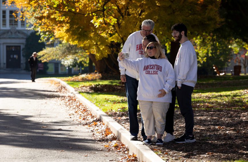 A family in matching Haverford College sweatshirts take a selfie under a tree full of golden fall leaves.