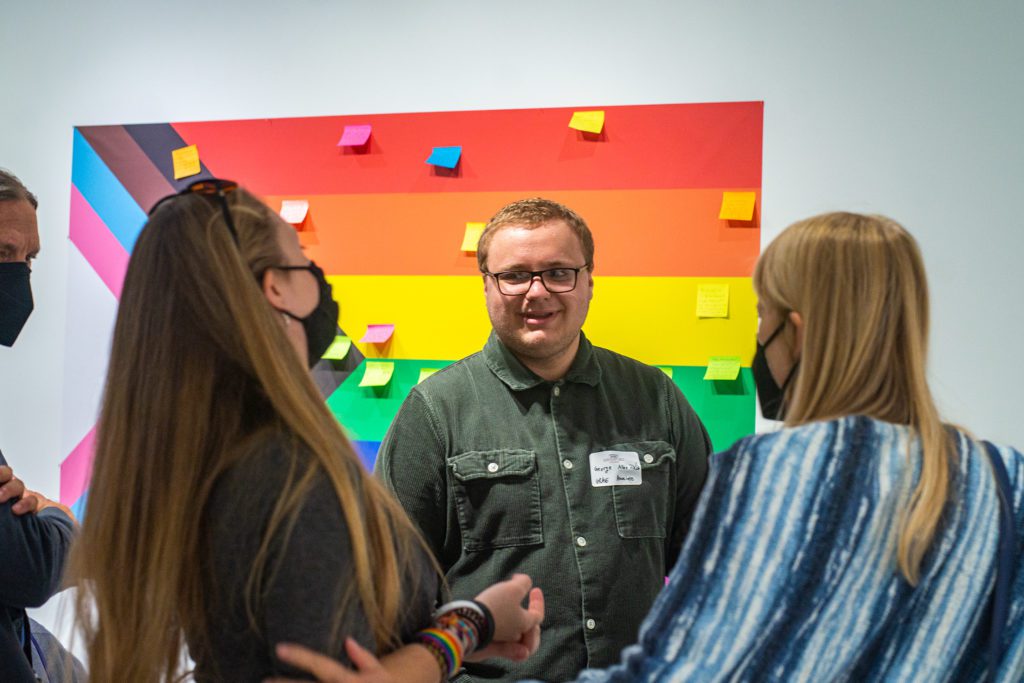 The three people stand in front of a large Pride flag covered in Post-It notes.