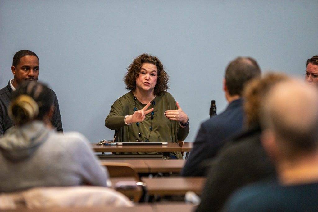 Barbara gestures with her hands while seated at a table at the front of the room.