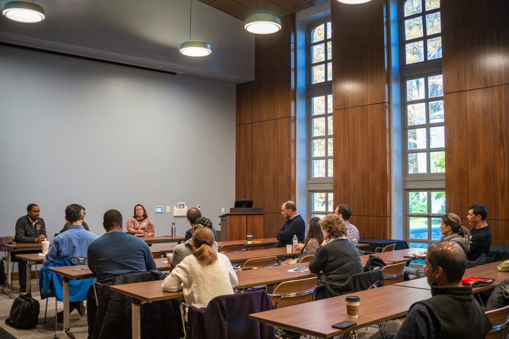 Deans sit at a long table in front of an audience seated at different long tables in the library.
