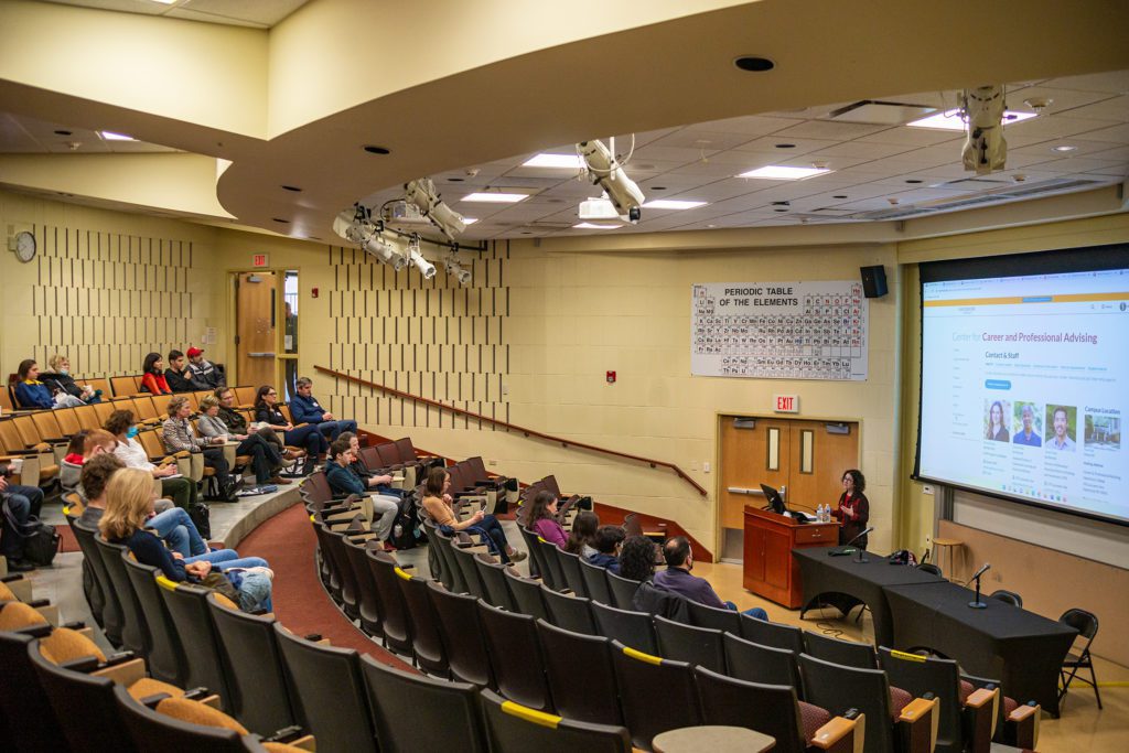 A woman speaks at a podium in a big lecture hall with a screen behind her showing all the staff of the Center for Career and Professional Advising.