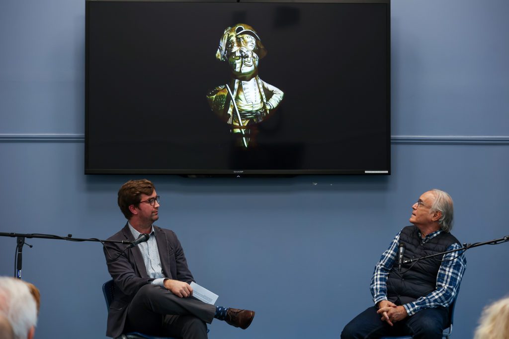 Two people seated at microphones under a screen with an image of George Washington on it.