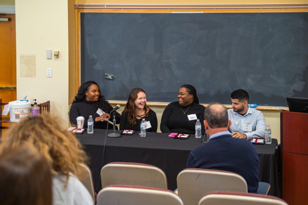 Four young alumni laugh, seated together at a table in front of microphones.