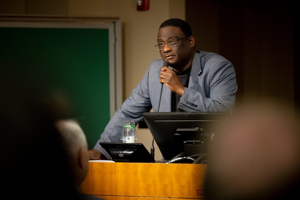Rel holds a microphone speaking at the podium.