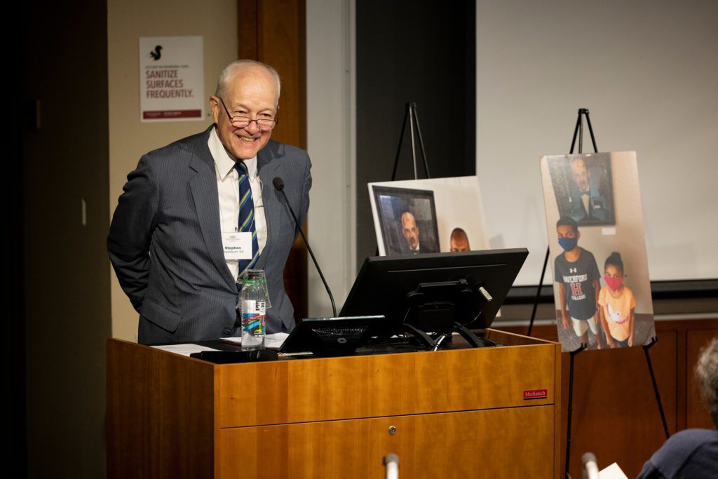 Stephen stands at a podium with photos taken with a painting of Ira Reid behind him.