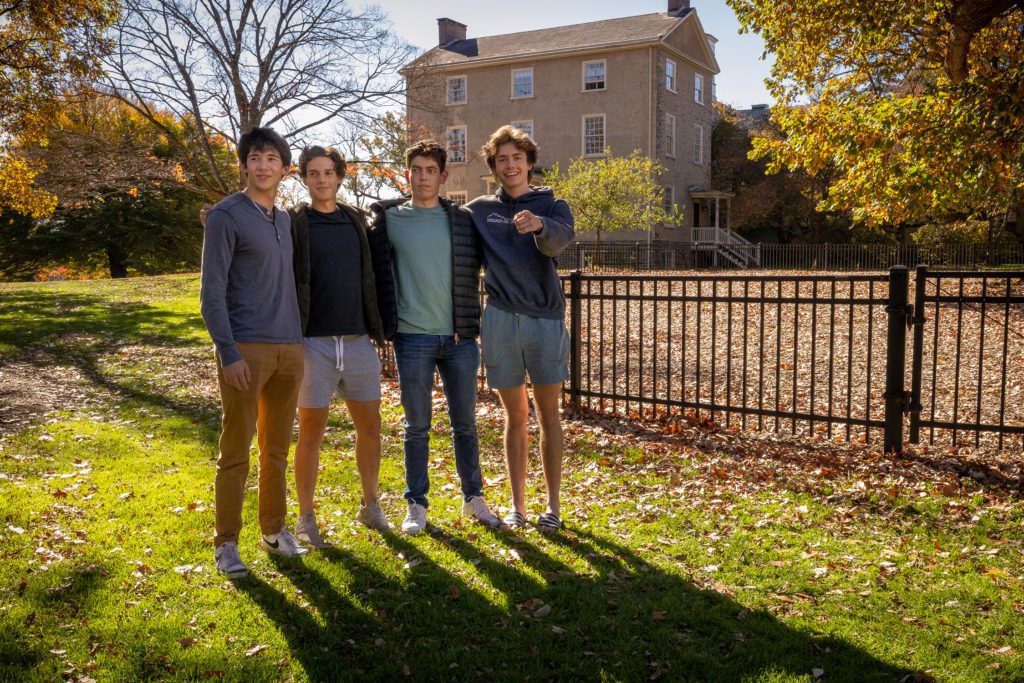 Four friends pose for a portrait with Founders Hall and fall foliage in the background.