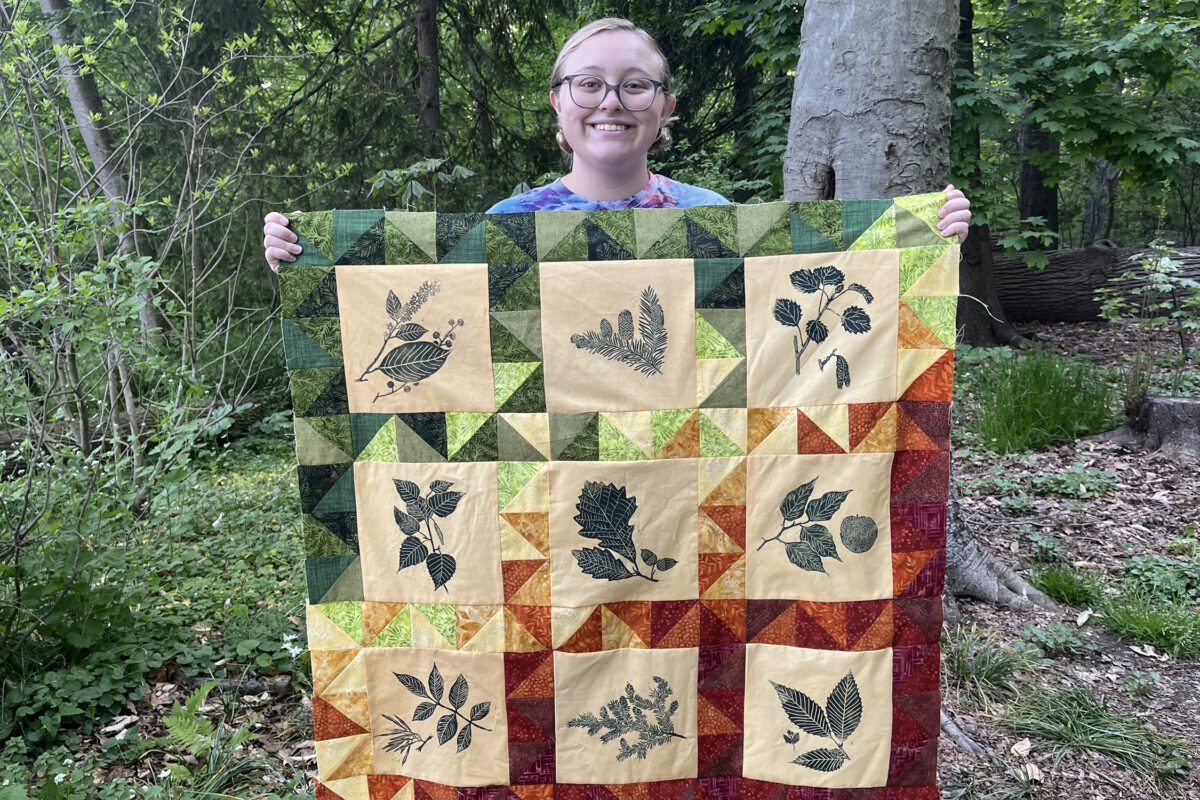 Sophia Wagner is surrounded by a natural setting. With two hands, she holds up a colorful quilt that features prints of native Pennsylvania plants.