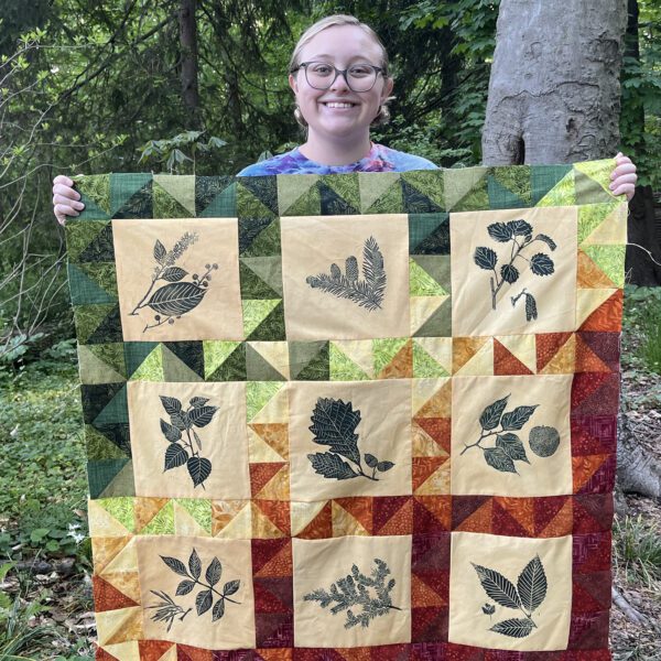 Sophia Wagner is surrounded by a natural setting. With two hands, she holds up a colorful quilt that features prints of native Pennsylvania plants.