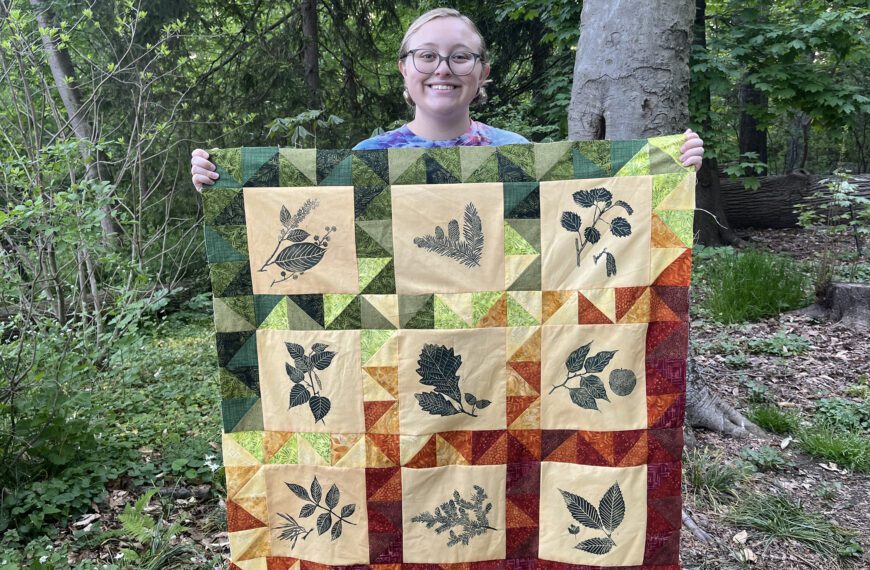 Sophia Wagner is surrounded by a natural setting. With two hands, she holds up a colorful quilt that features prints of native Pennsylvania plants.