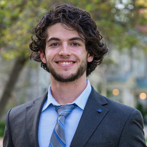 Wearing a dark suit jacket, light blue shirt, and a blue striped tie, Lucas Winkler is pictured in a portrait on Haverford's campus.