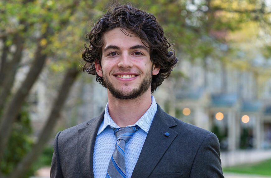 Wearing a dark suit jacket, light blue shirt, and a blue striped tie, Lucas Winkler is pictured in a portrait on Haverford's campus.
