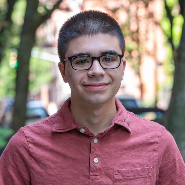 On a city street, Theo Schefer is pictured in a portrait. He wears a light red shirt with three buttons and dark glasses.