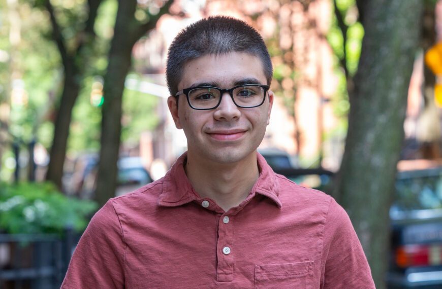 On a city street, Theo Schefer is pictured in a portrait. He wears a light red shirt with three buttons and dark glasses.