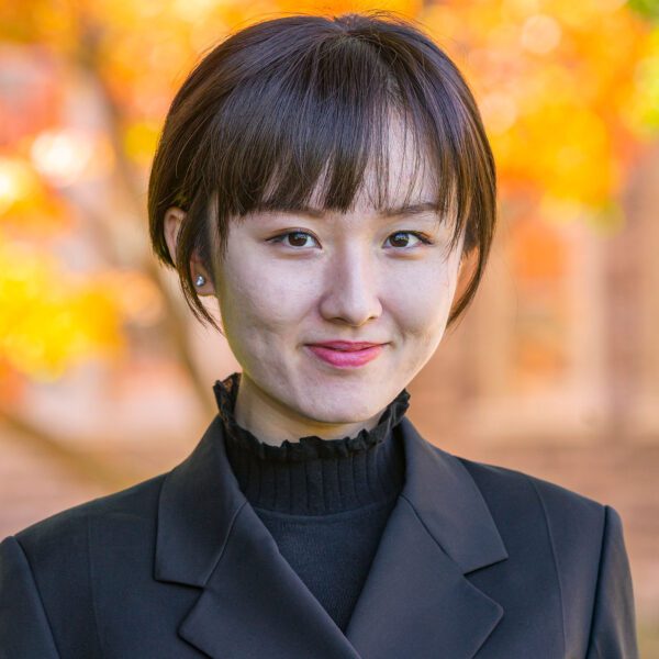 Maggie Zhang is pictured in a portrait on Haverford's campus. She wears a dark shirt and jacket against fall foliage.