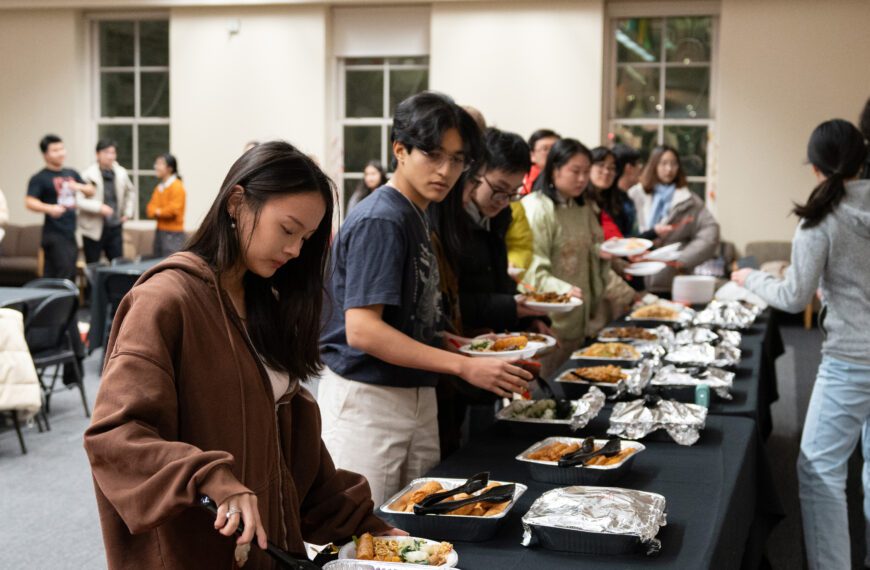 Students stand in a line to serve themselves food from a buffet lined up on several tables.