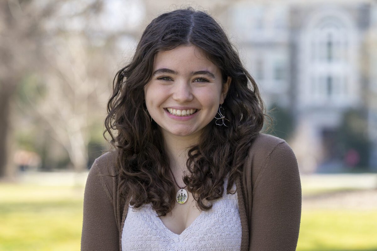 A young woman with long curly brown hair smiles at the camera while standing outdoors on a sunny day. She is wearing a light knit top, a brown cardigan, silver earrings, and a pendant necklace with a tree design. Blurred trees and a stone building are visible in the background.