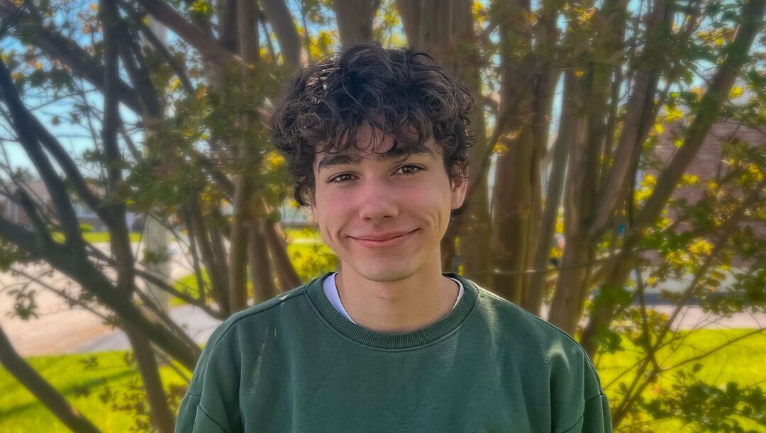 A young man with curly dark hair and dimples smiles gently at the camera. He is wearing a dark green crewneck sweatshirt and standing outdoors in front of a tree with thin branches and green leaves. The background is sunlit and slightly blurred, suggesting a warm, pleasant day.