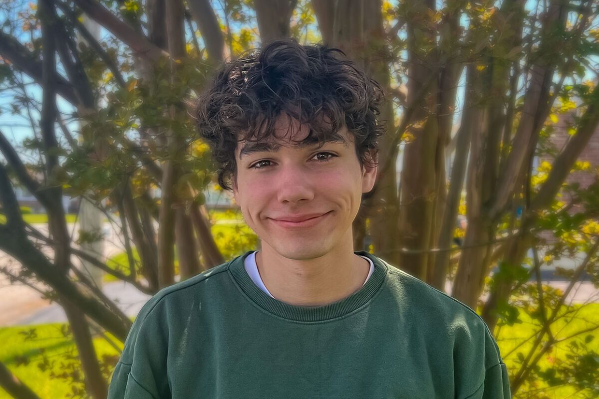 A young man with curly dark hair and dimples smiles gently at the camera. He is wearing a dark green crewneck sweatshirt and standing outdoors in front of a tree with thin branches and green leaves. The background is sunlit and slightly blurred, suggesting a warm, pleasant day.