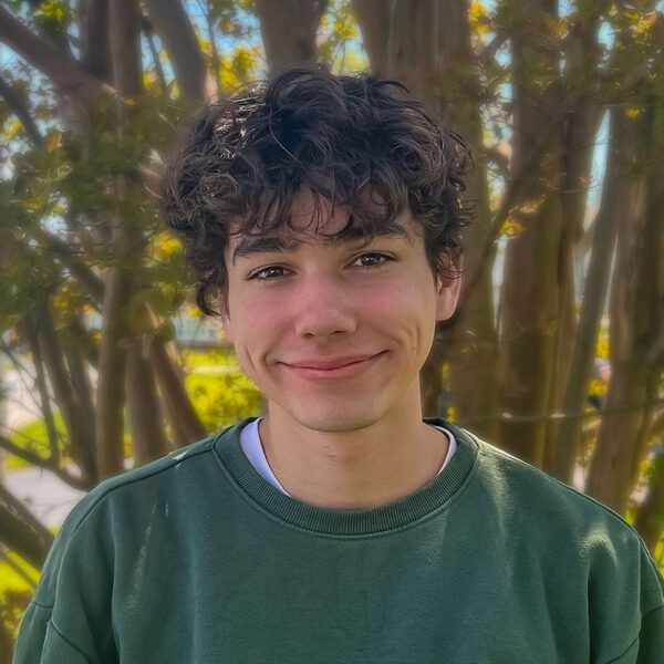 A young man with curly dark hair and dimples smiles gently at the camera. He is wearing a dark green crewneck sweatshirt and standing outdoors in front of a tree with thin branches and green leaves. The background is sunlit and slightly blurred, suggesting a warm, pleasant day.