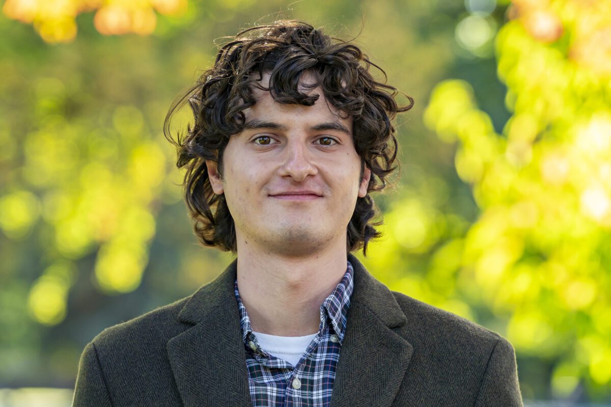 A young man with curly brown hair, wearing a plaid shirt and dark blazer, stands outdoors in front of a background of soft-focus green and yellow foliage.