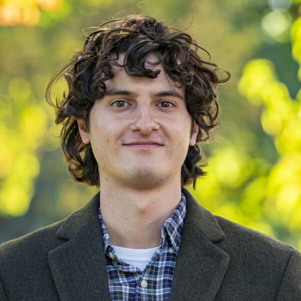 A young man with curly brown hair, wearing a plaid shirt and dark blazer, stands outdoors in front of a background of soft-focus green and yellow foliage.
