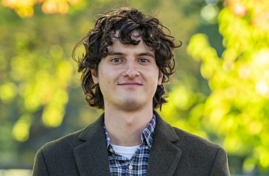 A young man with curly brown hair, wearing a plaid shirt and dark blazer, stands outdoors in front of a background of soft-focus green and yellow foliage.