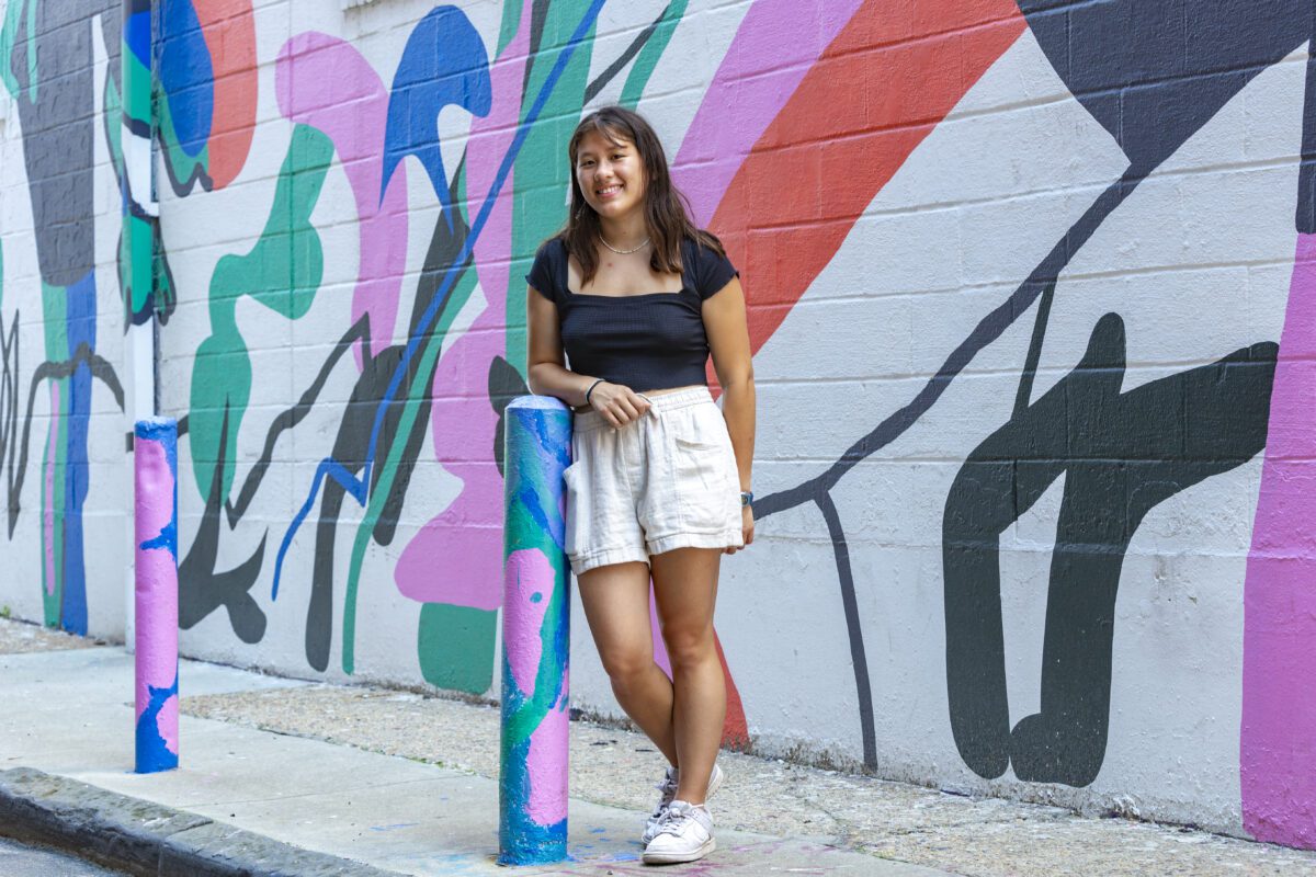 Kira Wu-Hacohen ’25 stands smiling in front of a colorful mural painted on a brick wall. She is wearing a black top, light shorts, and white sneakers, and is leaning casually against a painted post on a city sidewalk.